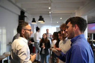 Two men talking in the foreground of a bright open room while other attendees mingle in small groups behind them