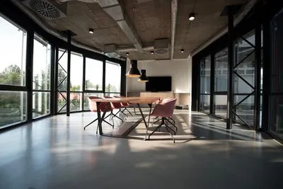 An empty meeting room with a long wooden table, chairs pulled up to it, and floor-to-ceiling windows on two sides