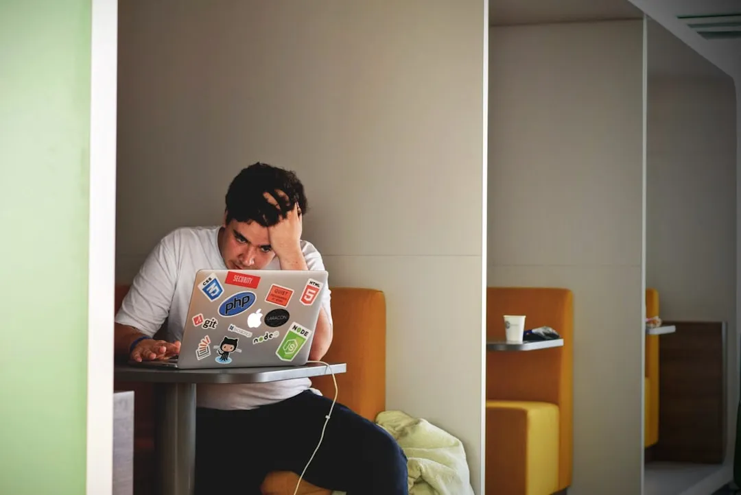 A frustrated man resting his head on his hand while looking at a laptop covered in tech stickers