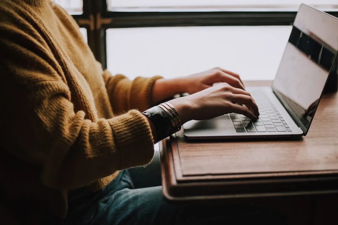 A person typing on a laptop keyboard by a train window, wearing a mustard sweater