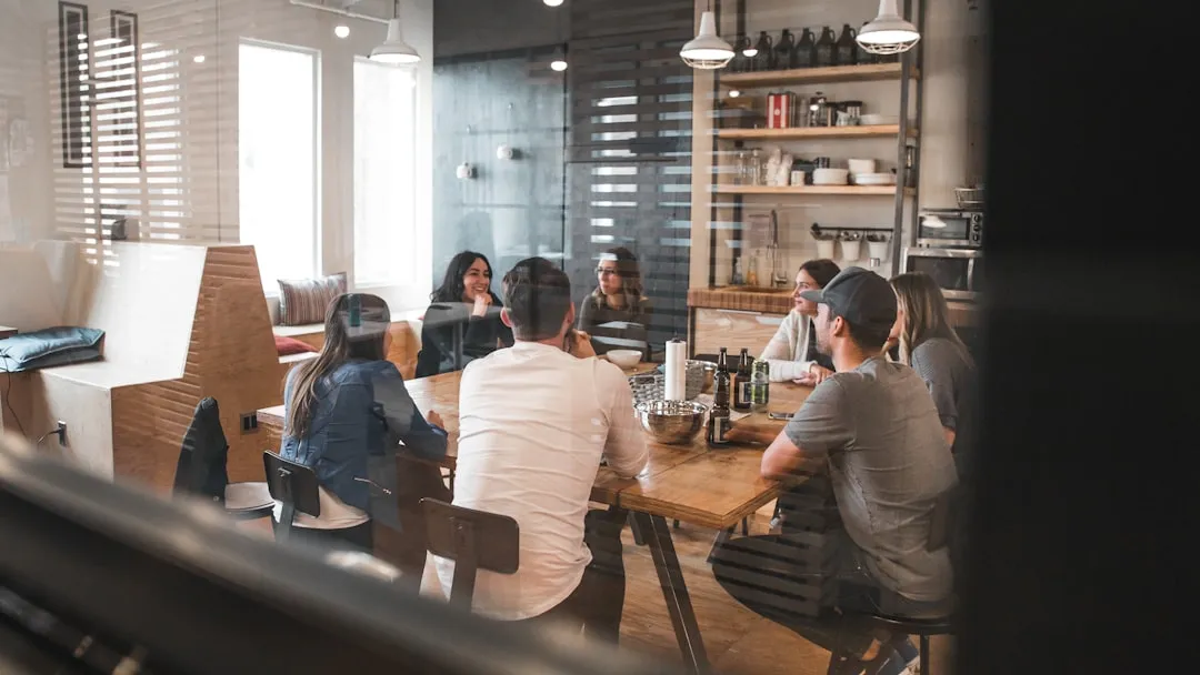 A group of coworkers gathered around a table in a casual office kitchen area, seen through glass