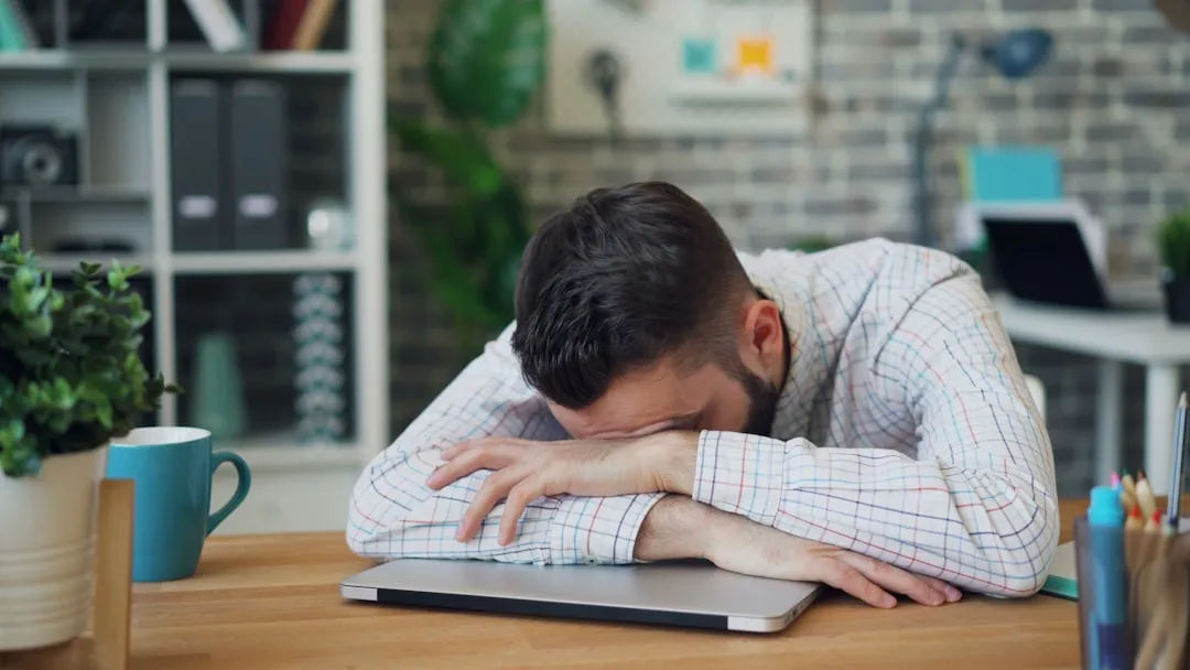 A man resting his head on his folded arms on a desk next to a closed laptop