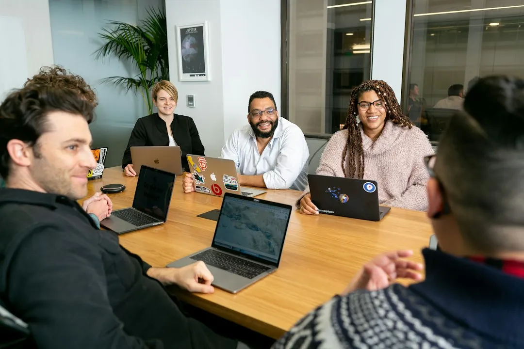 Four people on laptops, two men and two women, listen to a colleague talking at a meeting table