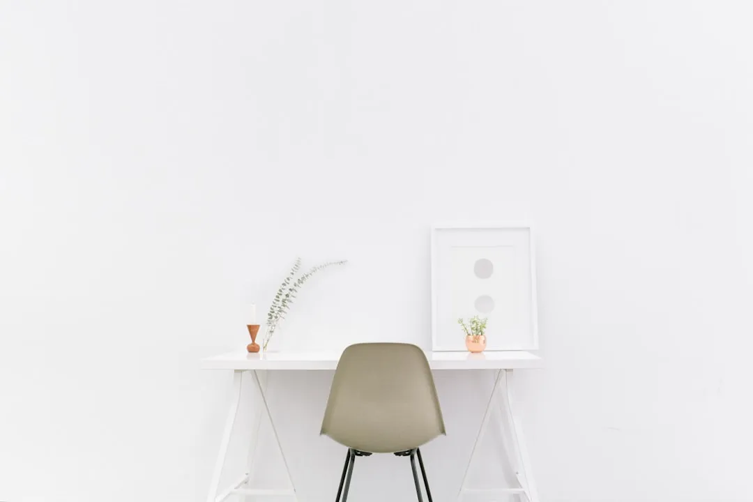 A minimalist white desk with a chair, framed art, and a small potted plant against a white wall